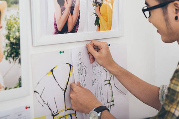 Young fashion designer pinning clothing sketches on wall in design studio, with fashion photography and mood board inspiration in background.