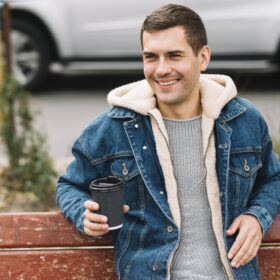 Man wearing a denim jacket with sherpa lining sitting on a bench and holding a coffee cup